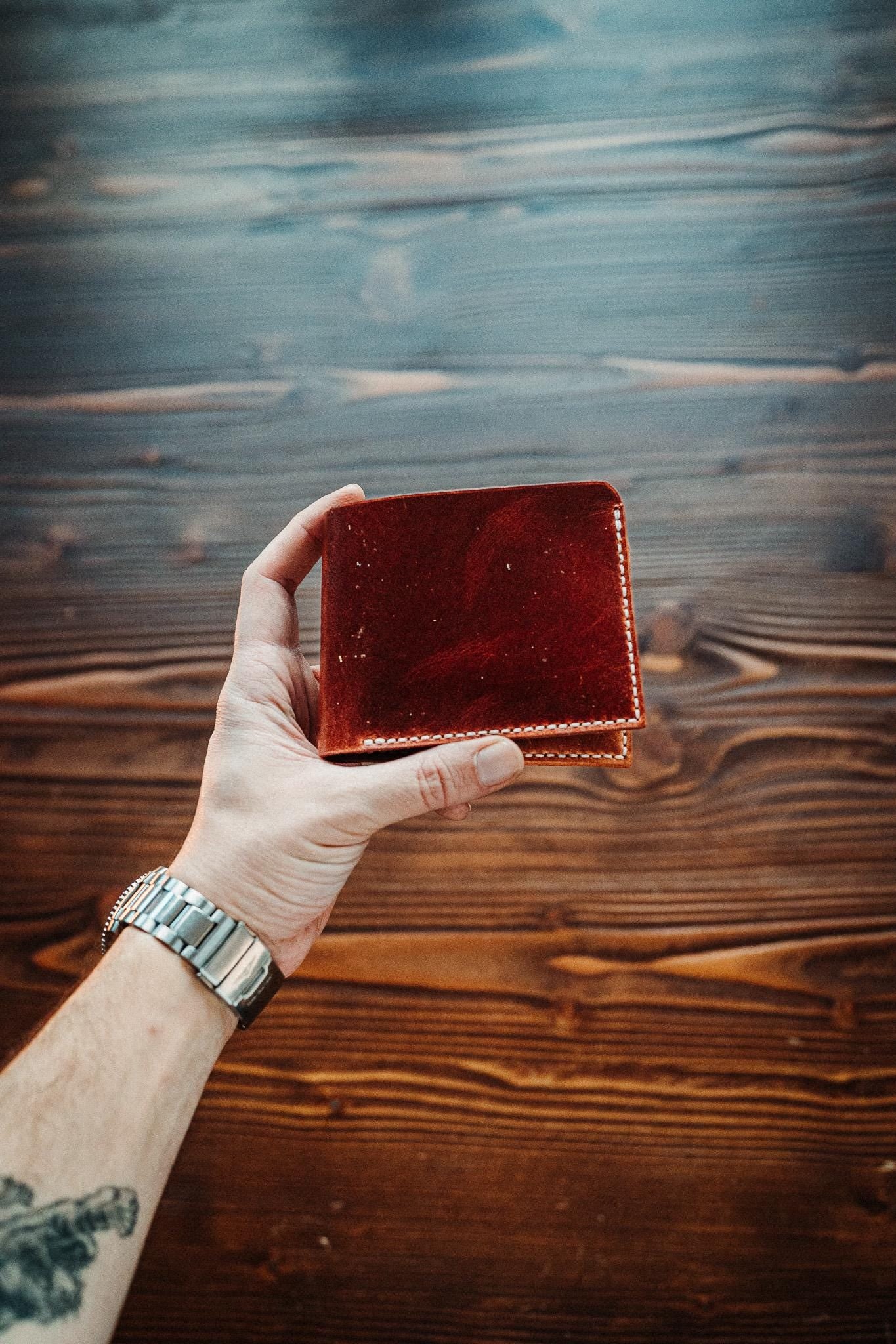 May include: A reddish-brown leather wallet with white stitching is held against a wooden background. The wallet is square and appears to be new. The background is a dark brown wood grain.